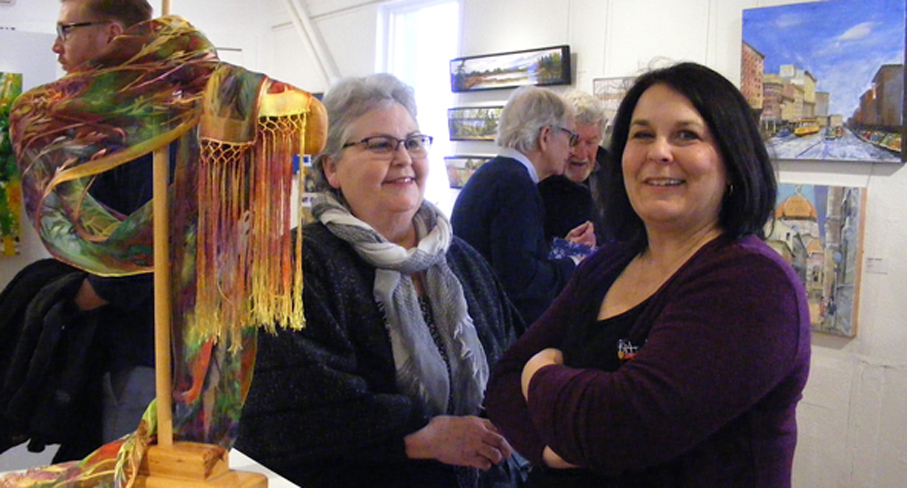 <p>Enjoying artistry – Michelle Sykes (left) from Ancaster and Picton artist Rollande Crawford stand next to a hand-painted silk scarf created by Amélie Koning de Bourbon during the opening reception at Arts on Main Saturday. The new “Spring Colour” show features works from more than 20 artists. (Terry McNamee/For The Gazette)</p>
