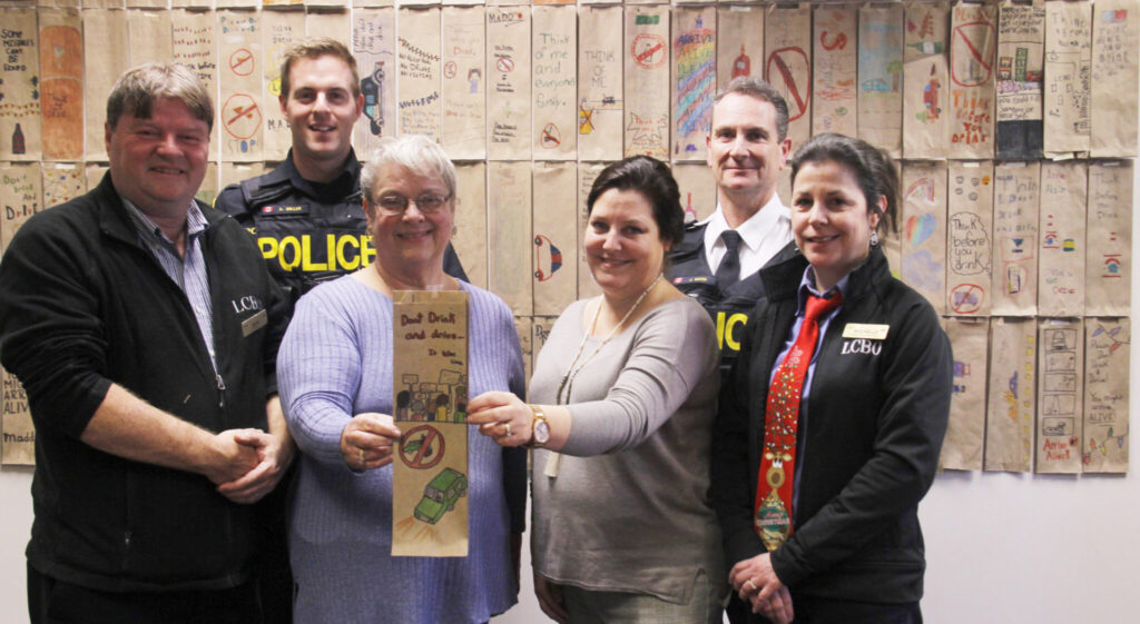 <p>ARRIVE ALIVE- Customers at local LCBO stores in Prince Edward County will be getting a gift this holiday season with their purchase as hand crafted designs by St. Greg’s students warning against the dangers of impaired driving will be part of their purchase. Back Row (from left) – Const. Aaron Miller and Staff-Sgt. John Hatch.<br />
In front are Scott Weir Picton LCBO manager, Gail McDougall of  St. Gregory Catholic School, Charlene Landry-Kyte St. Gregory Catholic School principal and Michelle Lavender of the LCBO. (Desirée Decoste/Gazette Staff)</p>
