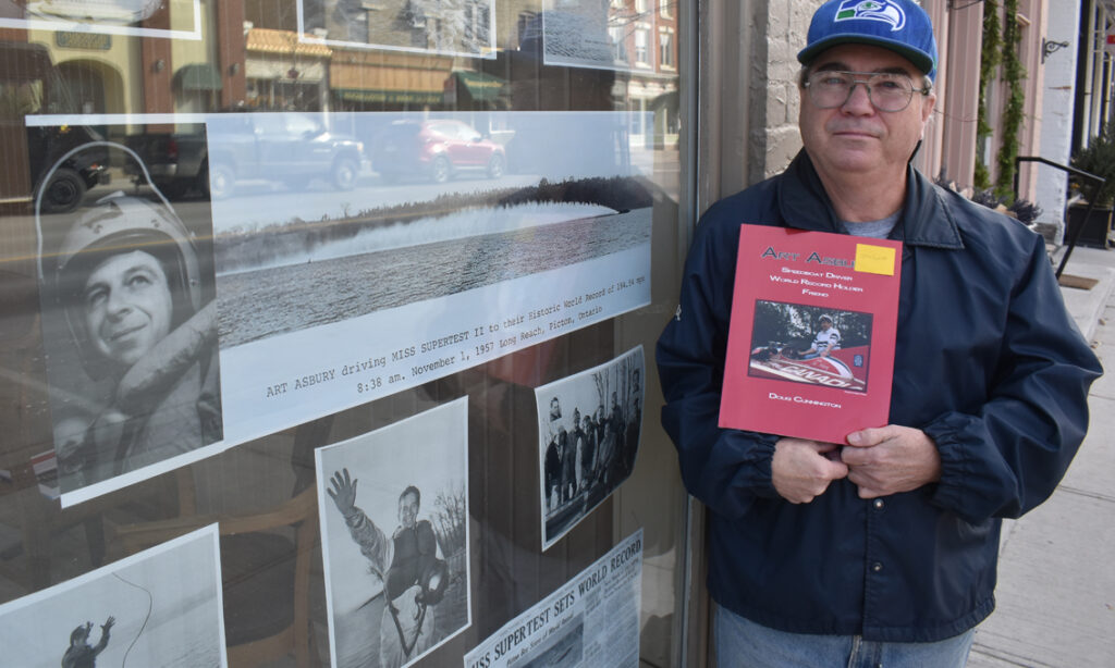 <p>MAKING THE MARK  Local powerboat historian John Lyons holds up Art Asbury: Speedboat Driver, World Record Holder, Friend by Doug Cunnington. The biography details Asbury’s Hall of Fame career including his run behind Miss Supertest II at Long Reach which set a world water speed record in 1957.  Lyons is in front of John’s Barbershop where a tribute to the world’s record attempt and Asbury hangs in the window. (Jason Parks/Gazette Staff)</p>
