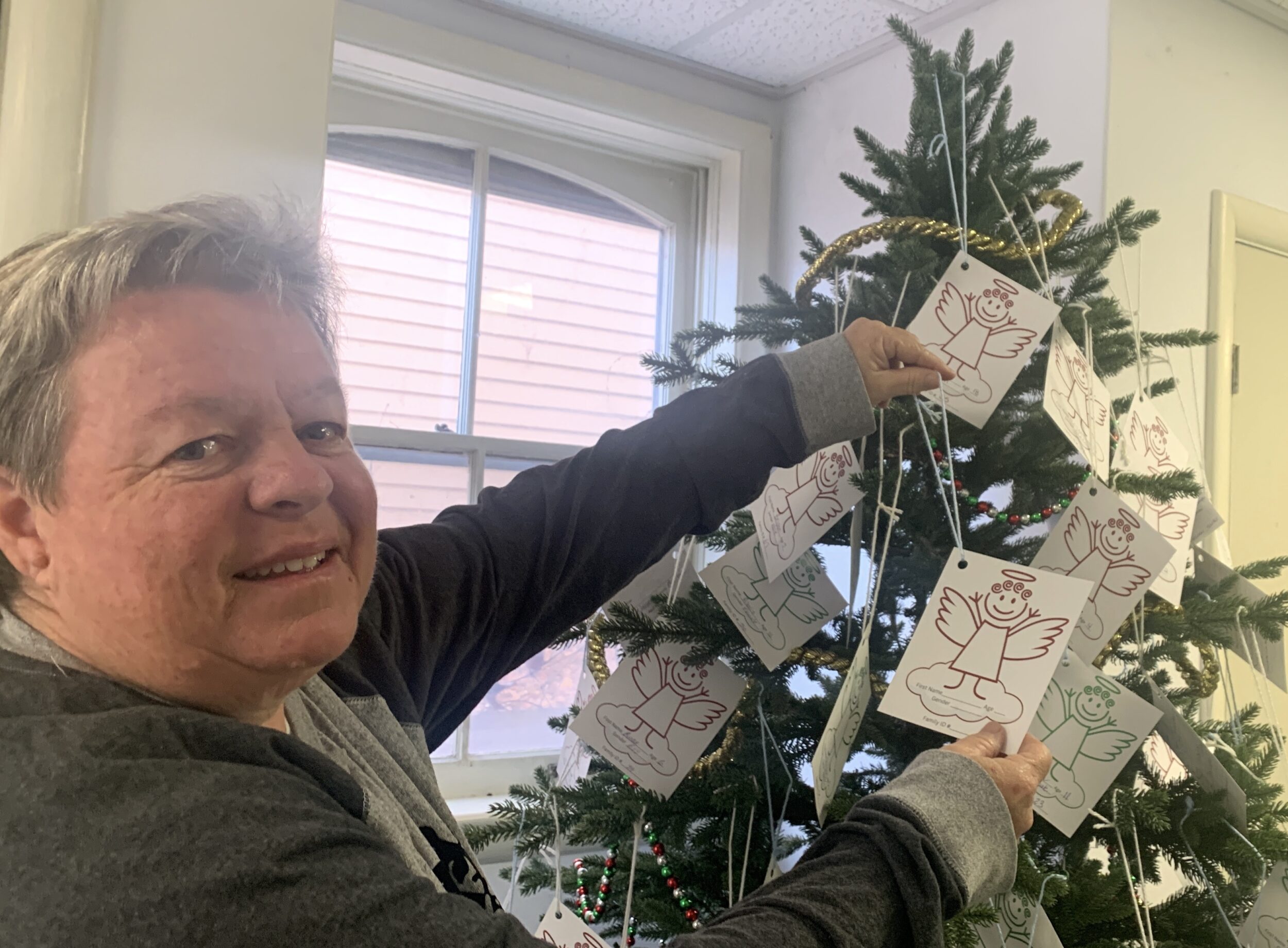 <p>Angel Tree coordinator Julie Miller hangs another paper angel on the Christmas Tree at the program office inside Saint Mary Magdalene’s Parish Hall. (Jason Parks/Gazette Staff)</p>

